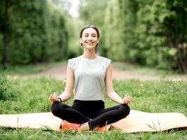 Eine lächelnde Frau sitzt im Schneidersitz auf ihrer Yoga-Matte, umgeben von Wiese und Bäumen.