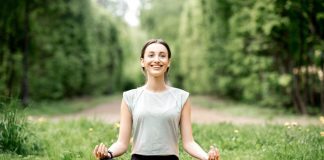 Eine lächelnde Frau sitzt im Schneidersitz auf ihrer Yoga-Matte, umgeben von Wiese und Bäumen.