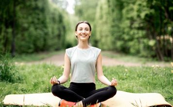 Eine lächelnde Frau sitzt im Schneidersitz auf ihrer Yoga-Matte, umgeben von Wiese und Bäumen.