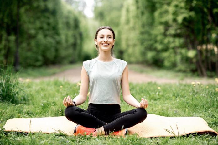 Eine lächelnde Frau sitzt im Schneidersitz auf ihrer Yoga-Matte, umgeben von Wiese und Bäumen.