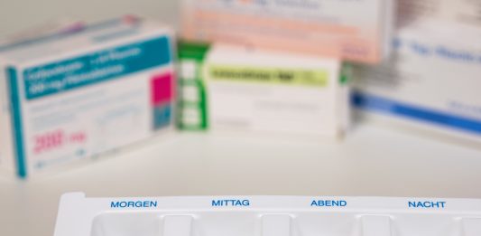 Medikamentenbox mit vier Fächern, beschriftet mit "Morgen", "Mittag", "Abend" und "Nacht", gefüllt mit verschiedenen Tabletten und Kapseln. Im Hintergrund unscharf Arzneimittelverpackungen. Ideal für die Organisation von Medikamenteneinnahmen.