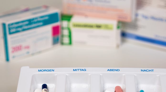 Medikamentenbox mit vier Fächern, beschriftet mit "Morgen", "Mittag", "Abend" und "Nacht", gefüllt mit verschiedenen Tabletten und Kapseln. Im Hintergrund unscharf Arzneimittelverpackungen. Ideal für die Organisation von Medikamenteneinnahmen.