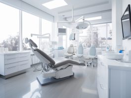 Modernes und helles Behandlungszimmer in einer Zahnarztpraxis mit dentaler Ausstattung, darunter ein Zahnarztstuhl, Leuchten, Monitore und medizinische Geräte. Große Fenster lassen viel Tageslicht herein.