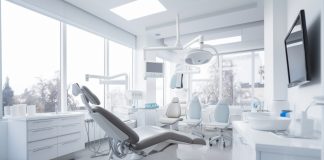 Modernes und helles Behandlungszimmer in einer Zahnarztpraxis mit dentaler Ausstattung, darunter ein Zahnarztstuhl, Leuchten, Monitore und medizinische Geräte. Große Fenster lassen viel Tageslicht herein.