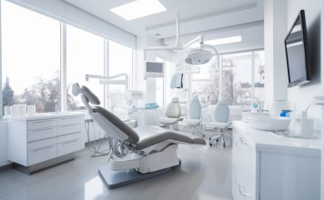 Modernes und helles Behandlungszimmer in einer Zahnarztpraxis mit dentaler Ausstattung, darunter ein Zahnarztstuhl, Leuchten, Monitore und medizinische Geräte. Große Fenster lassen viel Tageslicht herein.