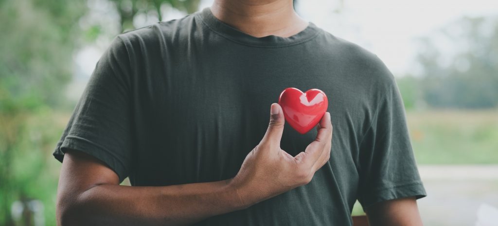 Ein Mann hält ein rotes Herzsymbol nahe seiner Brust, das die Bedeutung von Herzgesundheit und regelmäßigen Gesundheitschecks für Männer symbolisiert. Im Hintergrund ist eine verschwommene Naturkulisse zu sehen.