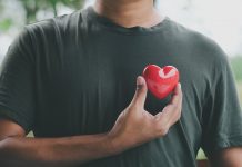 Ein Mann hält ein rotes Herzsymbol nahe seiner Brust, das die Bedeutung von Herzgesundheit und regelmäßigen Gesundheitschecks für Männer symbolisiert. Im Hintergrund ist eine verschwommene Naturkulisse zu sehen.