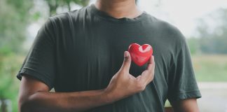 Ein Mann hält ein rotes Herzsymbol nahe seiner Brust, das die Bedeutung von Herzgesundheit und regelmäßigen Gesundheitschecks für Männer symbolisiert. Im Hintergrund ist eine verschwommene Naturkulisse zu sehen.