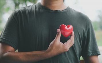 Ein Mann hält ein rotes Herzsymbol nahe seiner Brust, das die Bedeutung von Herzgesundheit und regelmäßigen Gesundheitschecks für Männer symbolisiert. Im Hintergrund ist eine verschwommene Naturkulisse zu sehen.