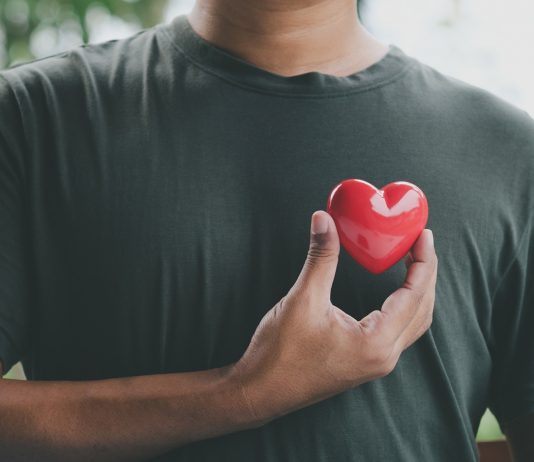 Ein Mann hält ein rotes Herzsymbol nahe seiner Brust, das die Bedeutung von Herzgesundheit und regelmäßigen Gesundheitschecks für Männer symbolisiert. Im Hintergrund ist eine verschwommene Naturkulisse zu sehen.