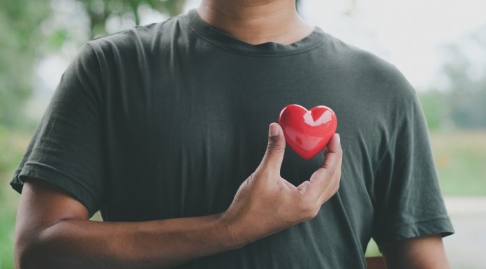 Ein Mann hält ein rotes Herzsymbol nahe seiner Brust, das die Bedeutung von Herzgesundheit und regelmäßigen Gesundheitschecks für Männer symbolisiert. Im Hintergrund ist eine verschwommene Naturkulisse zu sehen.